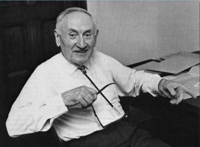 Seated, smiling old man in white shirt sitting down pointing at something.