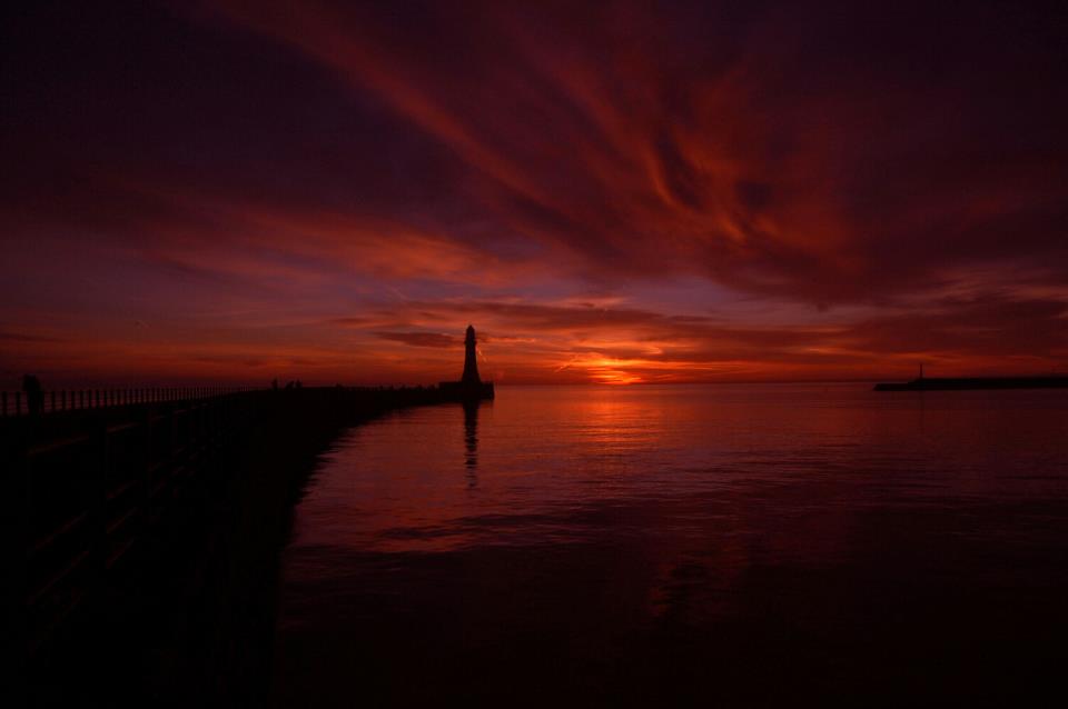Almanacs: Deep red sunset over a sea inlet with a distant silhouetted lighthouse.