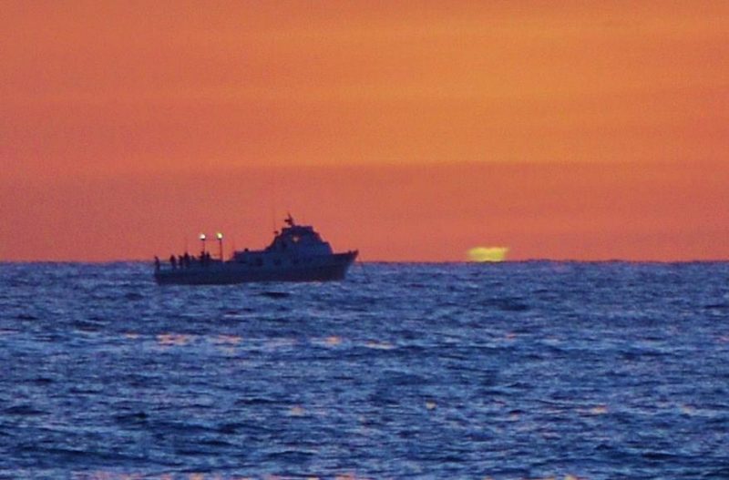Orange sky, blue sea, tugboat silhouette with green smudge on horizon.