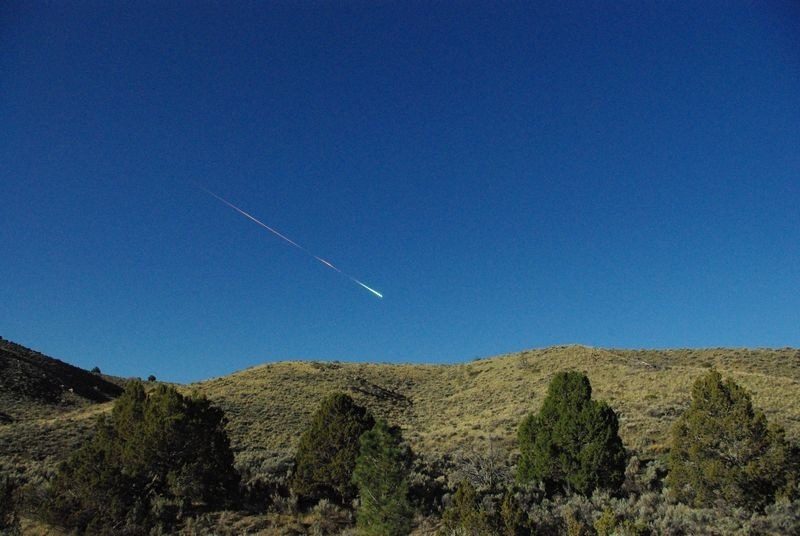 Bright meteor in blue sky above hills and trees.
