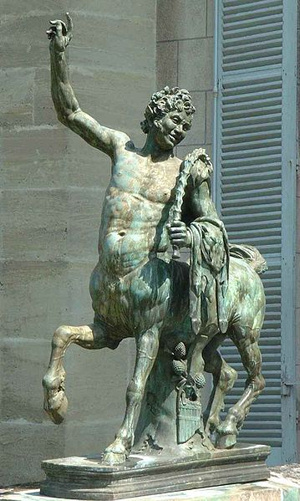 Greenish bronze statue of a centaur with the body and face of a young man, holding his right arm up.