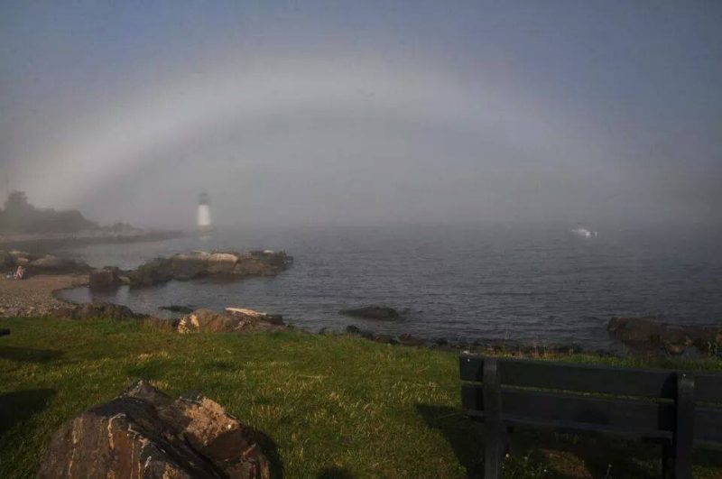 Diffuse white arc over rocky seacoast with white lighthouse in distance.