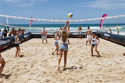People with short shadows playing volleyball in a sandy court.