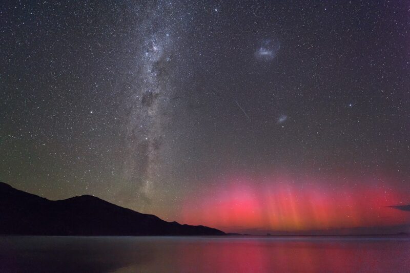Magellanic Clouds: Red aurora on the horizon, fuzzy white band of the Milky Way, and two small, fuzzy, glowing ovals.