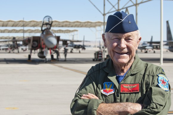 Old man in flight uniform standing in front of distant fighter plane.