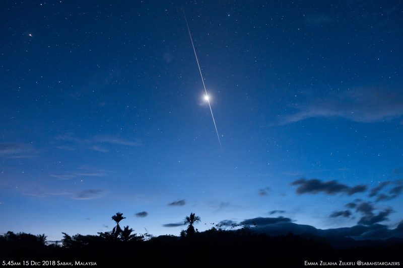 Meteors: Bright light in middle of elongated flash in dark blue sky with small clouds and stars.