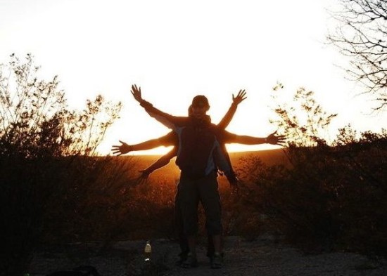 People in field standing behind each other with arms outstretched with sun behind them.