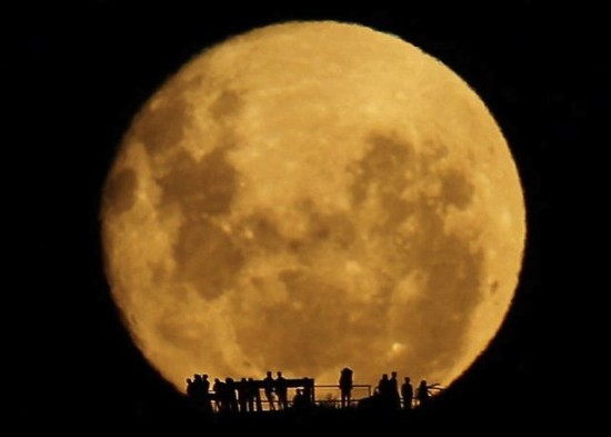 There is a big orange moon and a dark sky. There are people's silhouettes at the bottom of the image, looking at the moon.