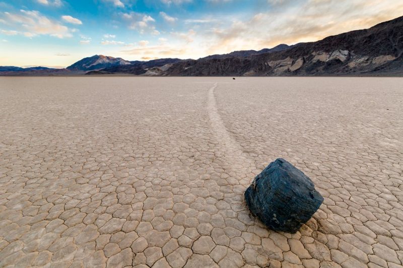 A faceted-looking black boulder with a track on a brown, cracked desert floor; mountains in background.