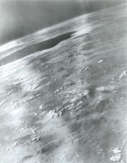 View of a portion of Earth from above, showing clouds and one dark patch. 