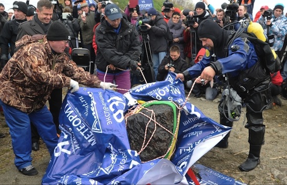 People standing around, two men tugging large tarp with big black rock on it.