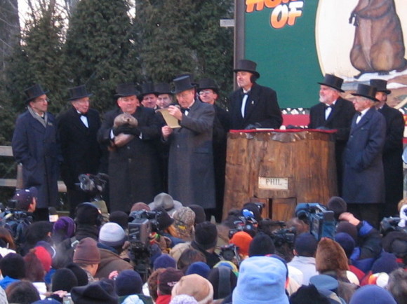 Men in dark winter coats and top hats, one holding a groundhog; crowd watching.