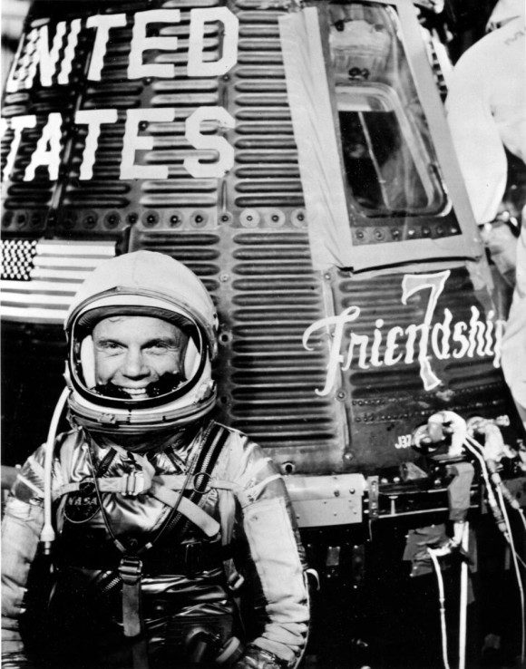 John Glenn: Smiling man in space suit standing next to a one-person space capsule.