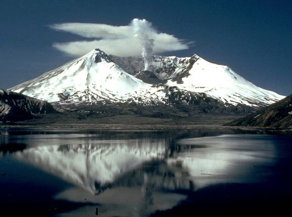 Sniegots kalns ar vulkāna krāteri virsotnē, dūmu plūmes centrā, mākoņi no aizmugures un atspulgs priekšējos ūdeņos.