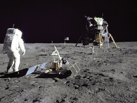 Astronaut in foreground with complicated device on the ground, lunar lander and flag in background.