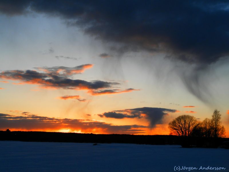 Sunset sky with horizontal clouds against orange, with curved virga streamers from dark cloud at top.