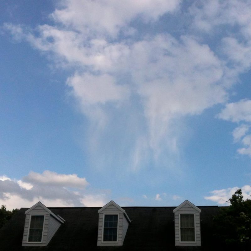 White fluffy clouds with white virga coming down from them, seen over a house rooftop.