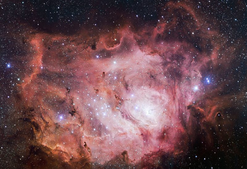The lagoon nebula: a celestial cloud of swirling gases, concentric pink ruffles containing patch of very bright stars.