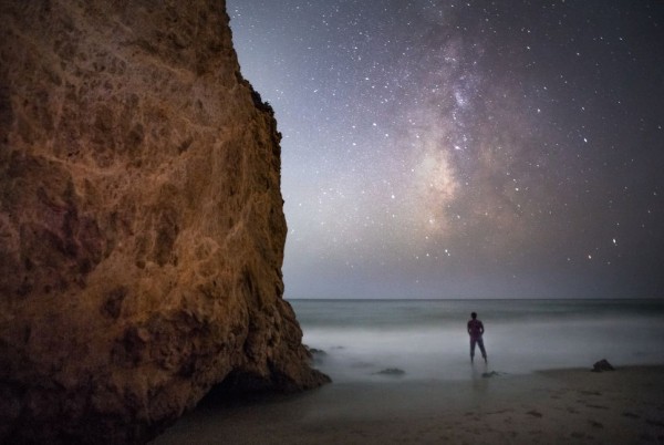 A man on a beach, next to a tall rocky cliff, under the Milky Way.