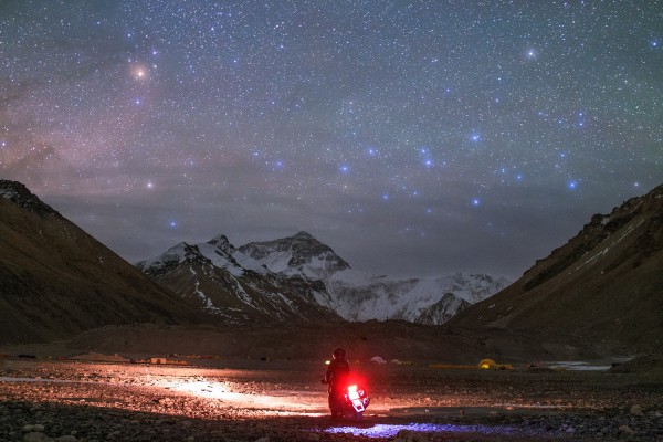 Riding the roof of the world. Everest Base Camp, Tibet, China. A lone motorcycle wends its way to Mount Everest's Base Camp, approaching from the Chinese side. In this darkening night sky, above the snow- and ice-flanked Himalayas, the yellow-red star Antares at the Scorpion's heart rises at left; to its right the stars of Centaurus shine their blue light over the top of the world. Mount Everest's name is Chomolungma in Tibetan language, often translated as 