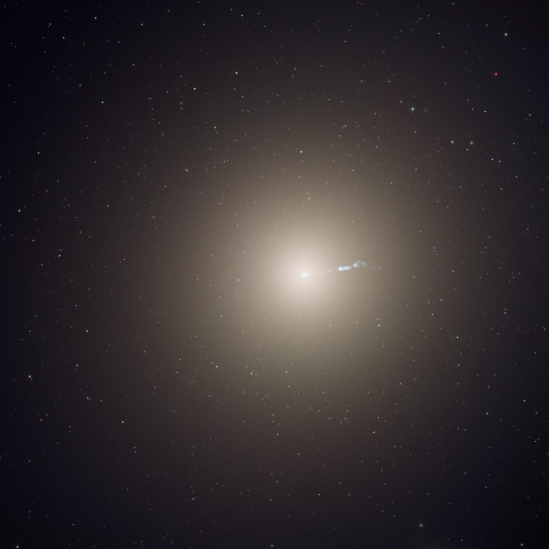 An elliptical galaxy shown as a central bright light with smoothly decreasing brightness the further from the center you go. From the center, a bluish light emanates towared the right. 