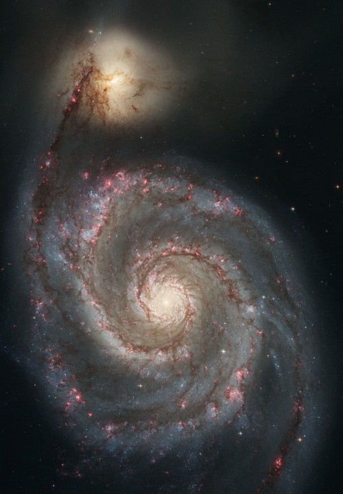 Galaxy with yellow-white center, two spiral arms dotted with pink areas. Bright yellow patch at end of one arm.