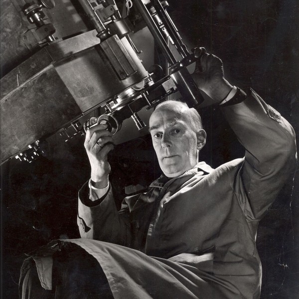Black-and-white image of a man at a telescope looking down at the camera.