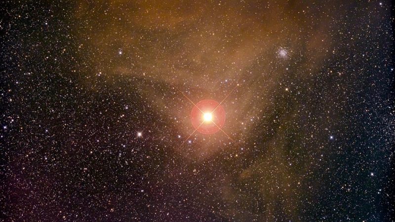 Star field with orange nebula, a bright orange star, Antares, and a small fuzzy round white object.