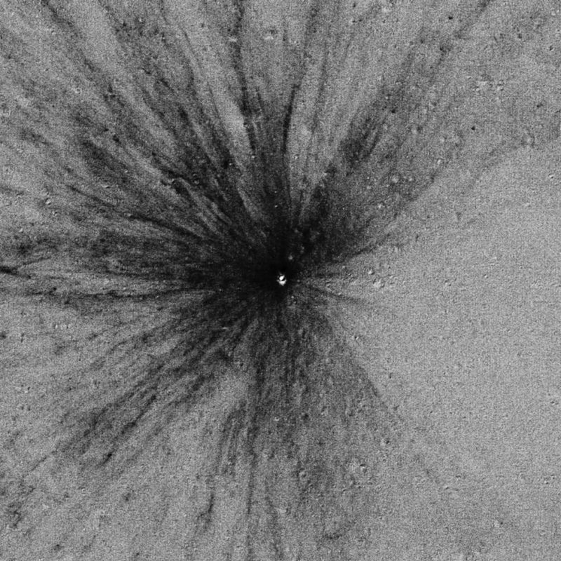 Temporal ratio image formed from two LROC Narrow Angle Camera images (after image divided by the before image) revealing a new 12 meter (~40 foot) diameter impact crater (Latitude: 36.536°N; Longitude: 12.379°E) formed between 25 October 2012 and 21 April 2013, scene is 1300 meters (~4200 feet) wide. New crater and its continuous ejecta are seen as the small bright area in the center, dark areas are the result of material blasted out of the crater to distances much further than previously thought. Image via NASA.