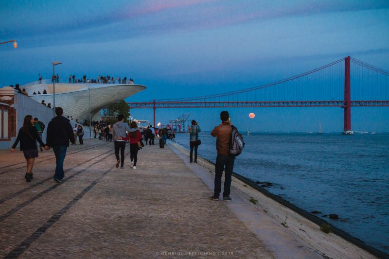 People standing on a modernistic building and oceanfront sidewalk watching moonrise under a distant bridge.