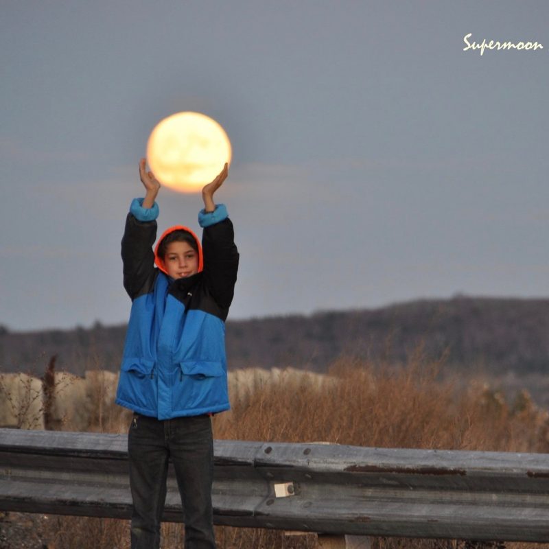 A boy in a jacket, outside, holding his hands up so that it looks like he's holding the full moon.