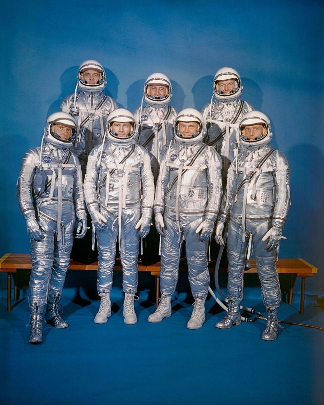 Group portrait of seven men in old-timey silver spacesuits, standing in two rows.