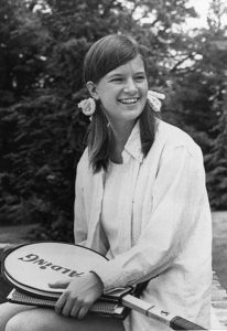 Black and white photo of smiling, seated, long-haired young woman holding a tennis racket.