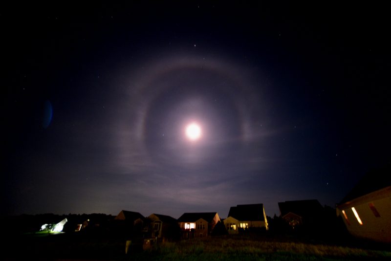 Overexposed moon with multiple light rings around it against dark sky, with housetops below.