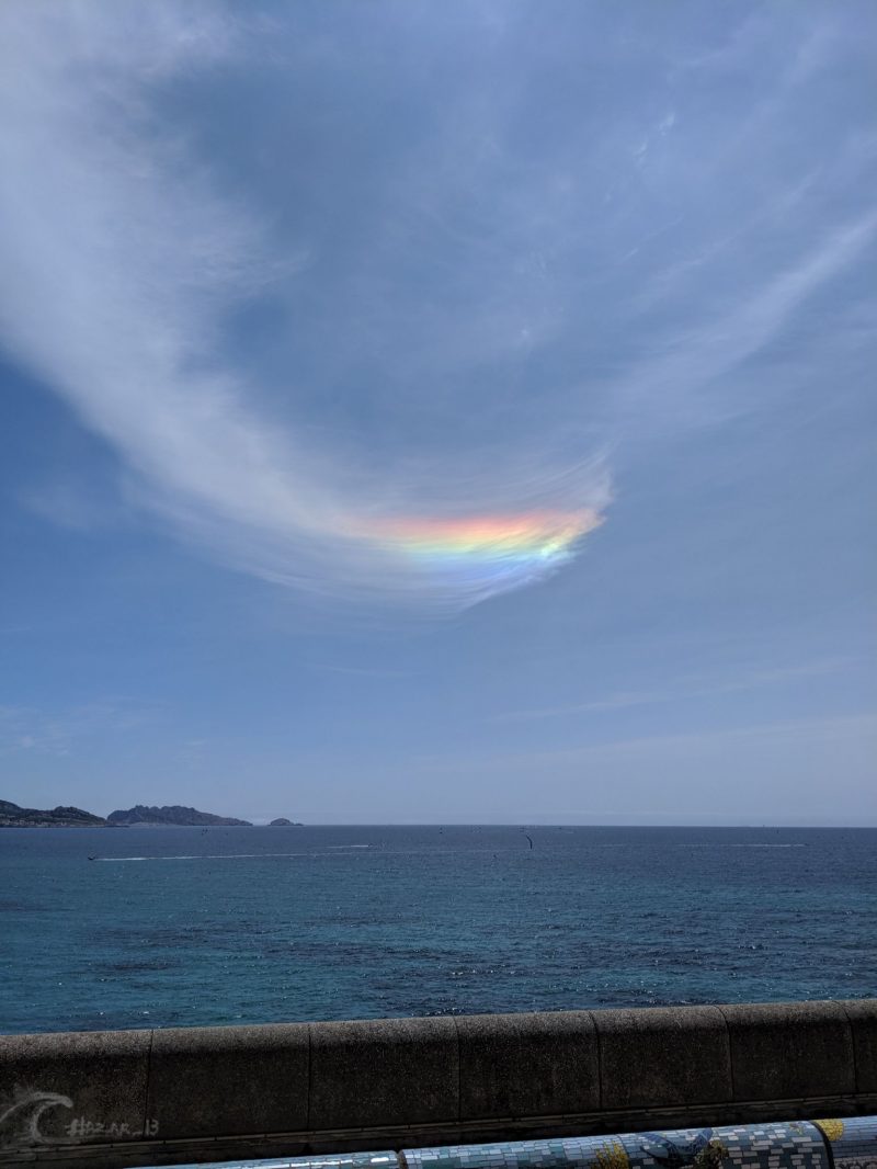 Wispy clouds over the sea. A line of rainbow colors in them, with blue on the bottom and red at top.
