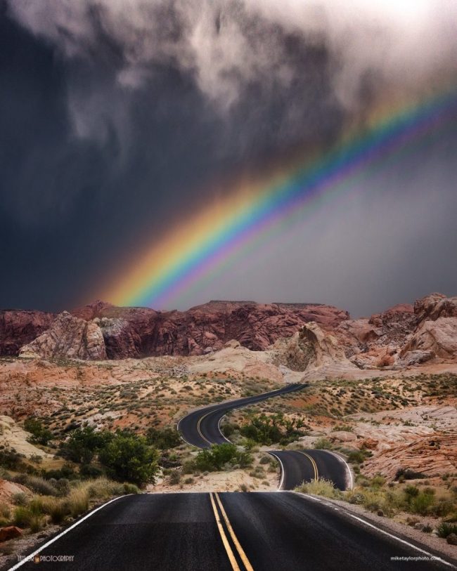 A rainbow with stormy clouds over badlands and a winding road.