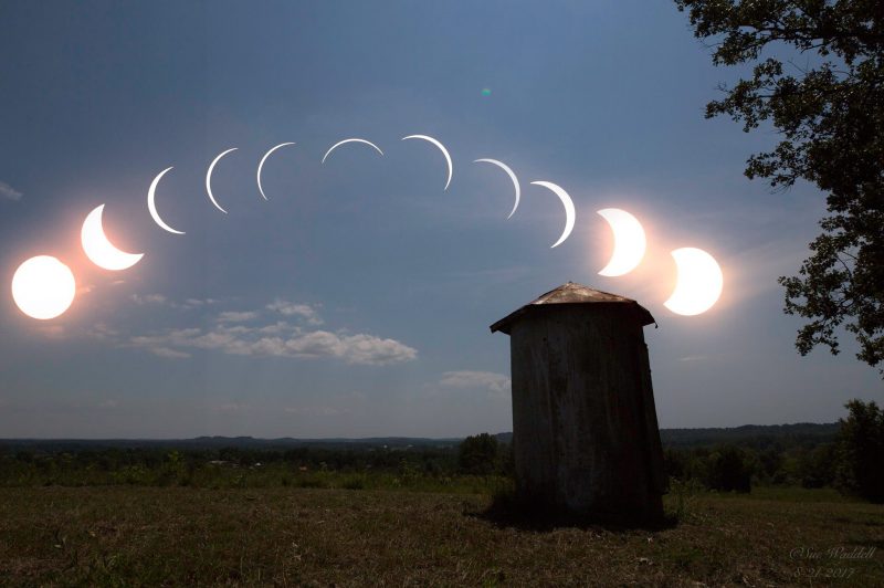 Arc of 11 crescent suns from wide to narrow to wide in sky above small outbuilding.