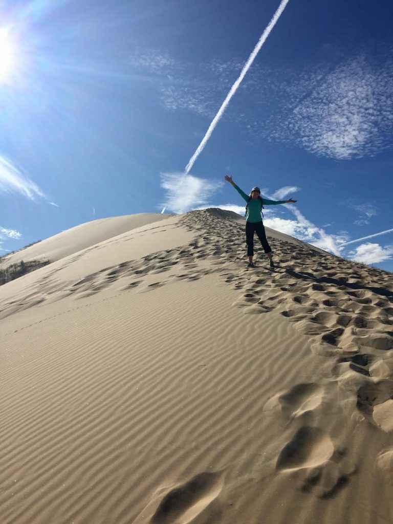 sand dune with person stretching up arms and diagonal jet contrail with a shadow above. 