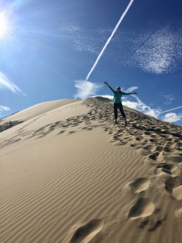 Top of sand dune with person stretching up arms and diagonal jet contrail with a shadow above.