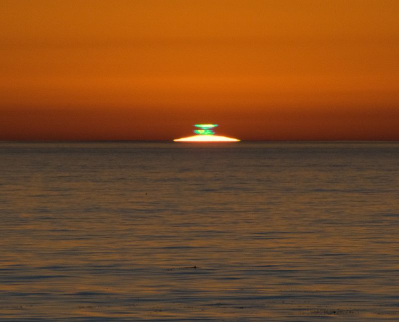Sliver of sun visible above ocean horizon with glowing green smudges above it.