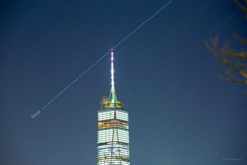 Line of small white dots across sky touching red light on top of brightly illuminated tower.