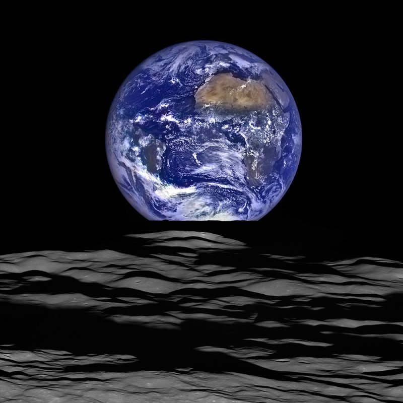 Full blue and white Earth viewed past hilly gray lunar landscape.