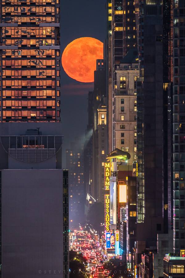 Large orange full moon between see-through glass skyscrapers above a lit-up city street.
