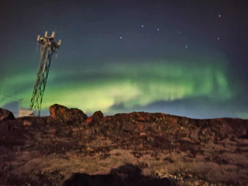 Big Dipper in autumn. Star pattern, with green curtains in the sky, behind rocky foreground with a radio tower.