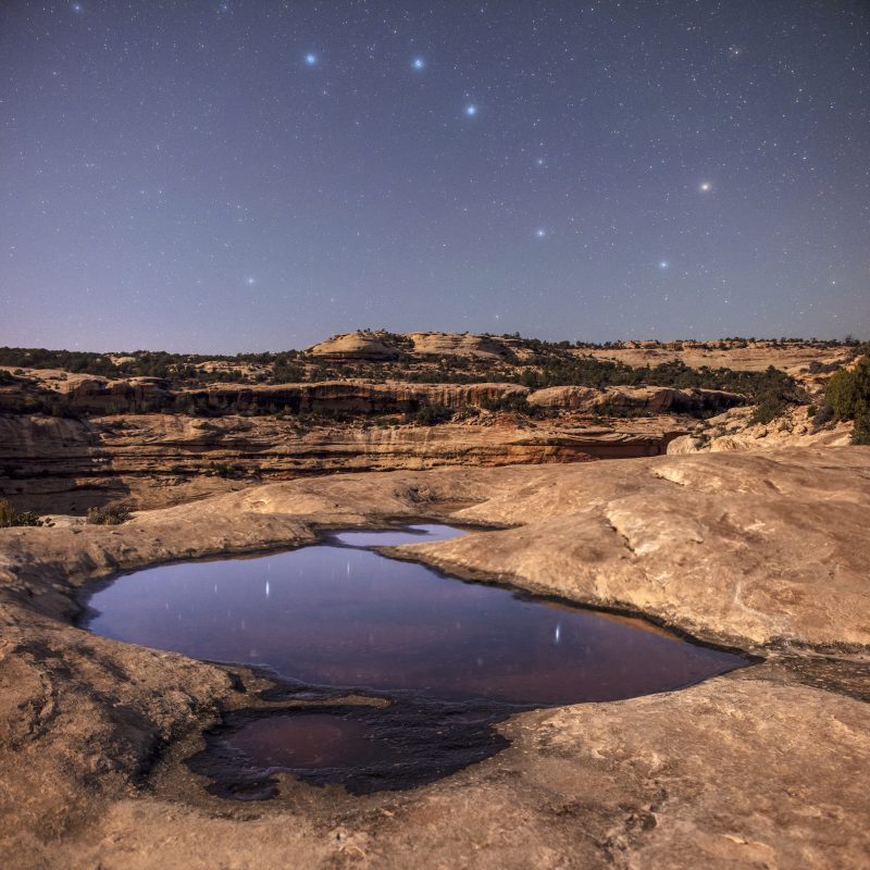 Desert scene with bright stars above and reflected in a pool among rocks and mesas.