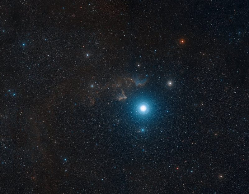 Dense star field with one very bright star and small, patchy cloudy area beside it.