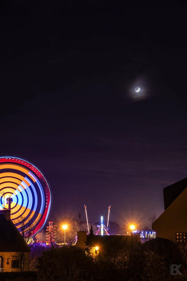 Night scene with large ferris wheel, with a crescent moon above it.