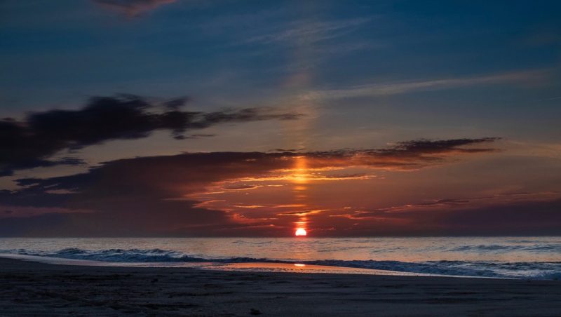 Orange sun rising over the ocean, with tall sun pillar above, brghter and dimmer as it passes through horizontal clouds.