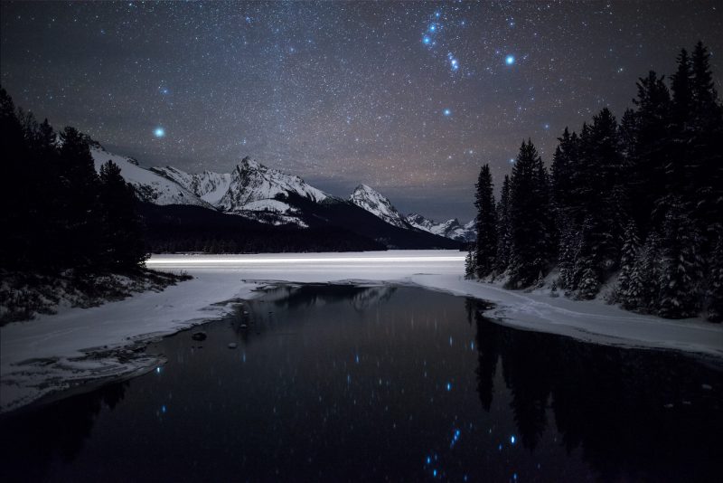 Starry sky, snowy mountains, black water with stars reflected in it.