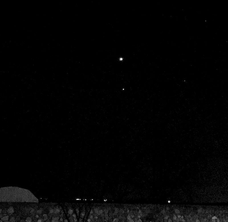 Two small round lights in a black sky seen over the top of a gray stone wall.